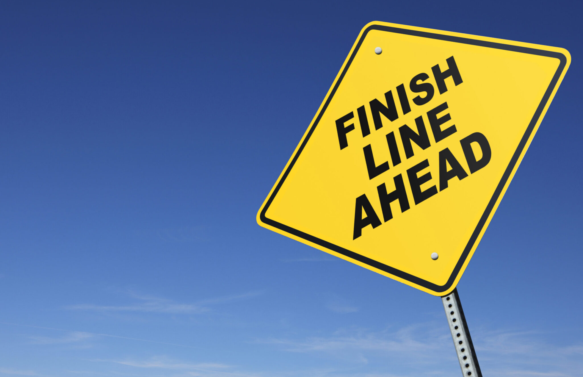 A  yellow road sign warning that the finish line is ahead.  A clear blue sky with wispy clouds at the horizon dominate the scene. Perspective is from a low camera angle. Road sign is placed on right side of image allowing for copy space on the left side of image.